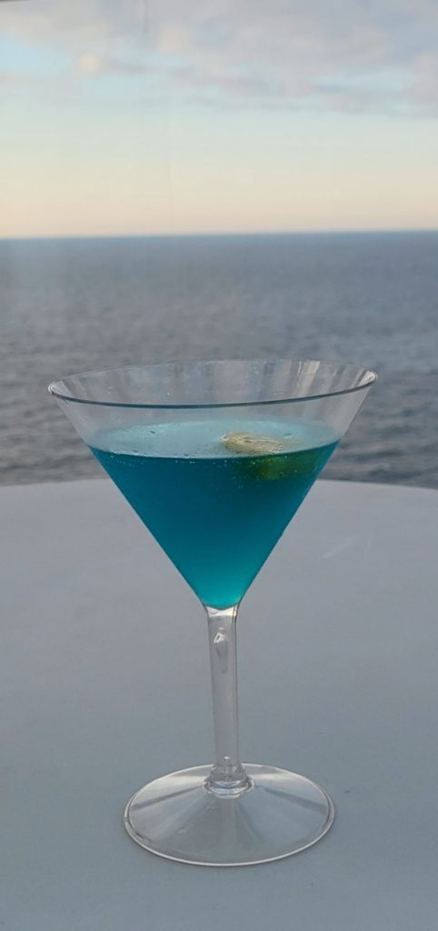 A blue cocktail in a martini glass with the ocean behind it.